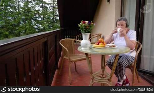 Pan of senior woman drinking her coffee and relaxing on terrace in summertime. Slow motion