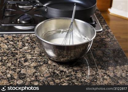Pancake batter in stainless steel bowl with whisk and stove top in background