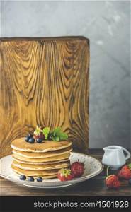 Pancake with srtawberry, blueberry and mint in ceramic dish, syrup from small ceramic jar on a light wooden table and gray background.