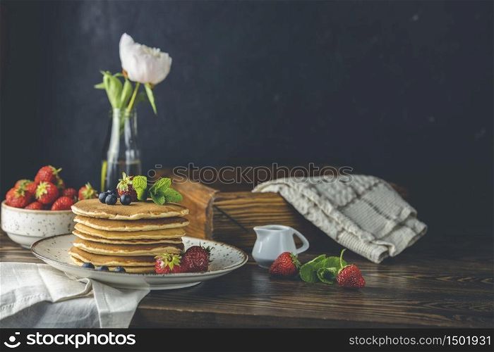 Pancake with srtawberry, blueberry and mint in ceramic dish, syrup from small ceramic jar and flowers on a dark wooden table and black background.