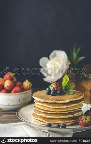 Pancake with srtawberry, blueberry and mint in ceramic dish, syrup from small ceramic jar and flowers on a dark wooden table and black background.