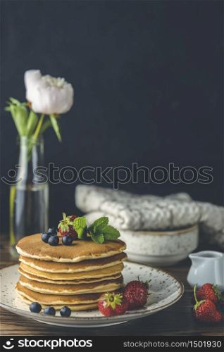 Pancake with strawberry, blueberry and mint in ceramic dish, syrup from small ceramic jar and flowers on a dark wooden table and black background.