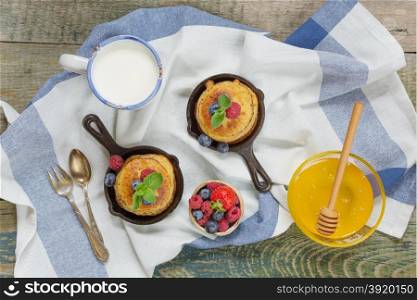 Pancakes in cast-iron frying pan with fresh berries and honey on the background of the old wooden boards and cup of milk