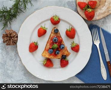 Pancakes with jam are located on a white plate, around the pancakes on a plate are fresh berries. Cutlery on a blue napkin. Breakfast, festive food. Gray background. View from above.