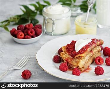 Pancakes with jam are located on a white plate, around the pancakes on a plate are fresh berries. Next to a glass of milk and sour cream. Breakfast Close-up. The table is decorated with an ivy branch.
