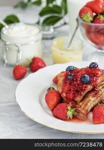 Pancakes with jam are located on a white plate, around the pancakes on a plate are fresh berries. Near jam and a cup of sour cream. Close-up. The table is decorated with an ivy branch.