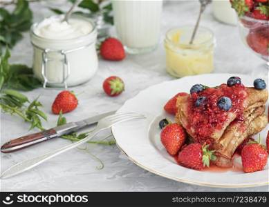 Pancakes with jam are located on a white plate, around the pancakes on a plate are fresh berries. Next to a glass of milk and sour cream. Breakfast Close-up. The table is decorated with an ivy branch.