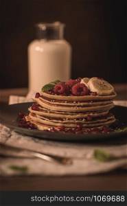 Pancakes with raspberries, banana slices, pomegranate seeds and honey on wooden vintage table.