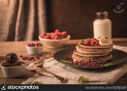 Pancakes with raspberries, banana slices, pomegranate seeds and honey on wooden vintage table.