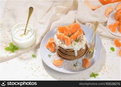 Pancakes with yogurt and tangerines on a light modern kitchen counter top with oat, thyme and mint leaves.