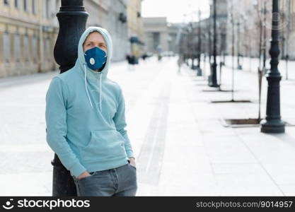 Pandemic coronavirus, Covid-19. Serious young man dressed in sweatshirt and jeans, wears respiratory mask to protect from virus or infection, walks through empty city streets during quarantine