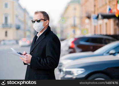 Pandemic coronavirus outbreak. Serious businessman poses outdoor near transport at street, holds modern cellular and newspaper, dressed in black coat, wears protective mask from coronavirus.