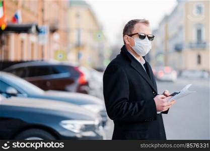 Pandemic coronavirus outbreak. Serious businessman poses outdoor near transport at street, holds modern cellular and newspaper, dressed in black coat, wears protective mask from coronavirus.