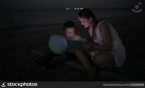 Panning shot of a little boy and his mom sitting by the sea with touchpad. Screen enlighting their faces