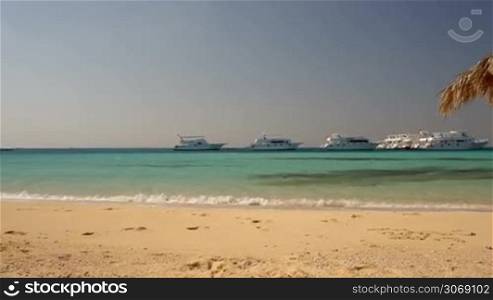 Panning shot of beautiful tropical scene with pure sea, straw sun umbrella and luxurious yachts in the distance