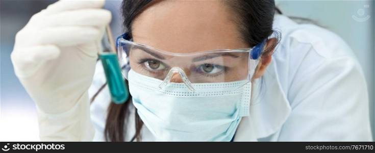 Panorama female medical or scientific researcher or doctor wearing a face mask looking at a test tube of liqud blue solution in a laboratory panoramic web banner header