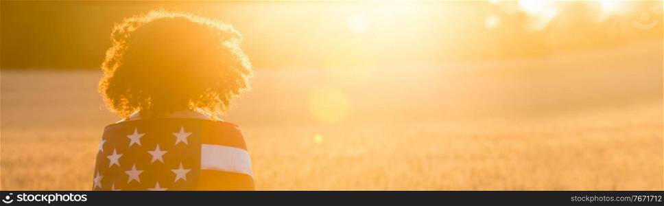 Panorama mixed race African American girl teenager female young woman in a field of wheat or barley crops wrapped in USA stars and stripes flag in golden sunset evening sunshine panoramic web banner