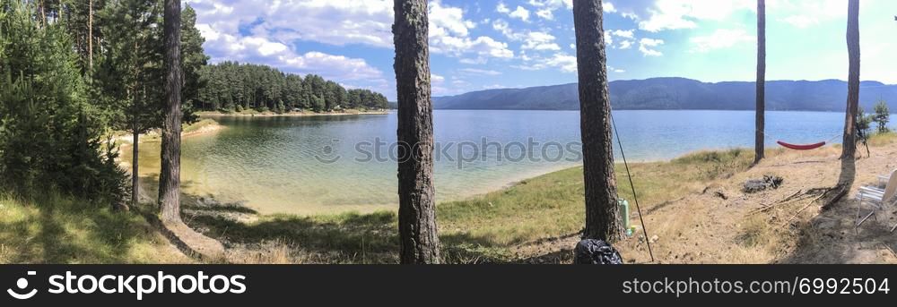 Panorama of forest and mountain lake. Hammock hooked on two pine trees. Sunny day. Outdoor panoramic concept.