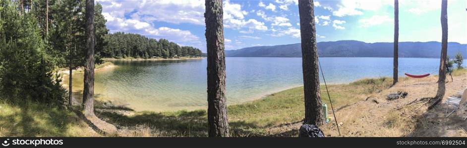 Panorama of forest and mountain lake. Hammock hooked on two pine trees. Sunny day. Outdoor panoramic concept.