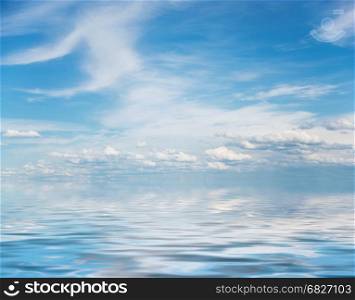 Panorama of vast blue summer sky with fluffy white cumulus and cirrus clouds reflected in a water surface with small waves