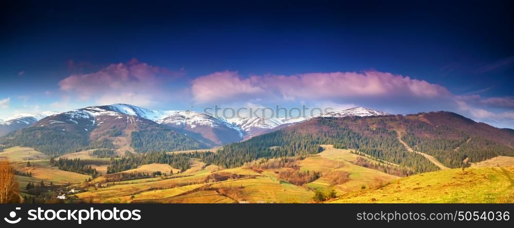 Panorama view of snow capped mountain peaks in spring time. Mountains in morning light. Deep blue sky. Idyllic Spring alpine morning landscape with fresh green meadows and snow-capped mountain tops in the background.
