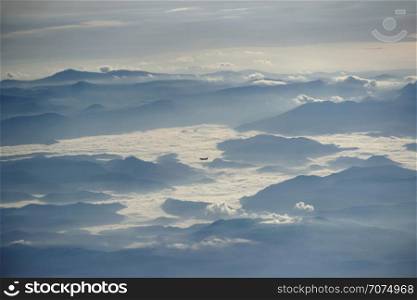 Panorama with flying plane high in sky above mountain ridge and clouds. Beautiful view from window of plane in evening. View from plane window. View from plane window to another plane flying over clouds and mountains
