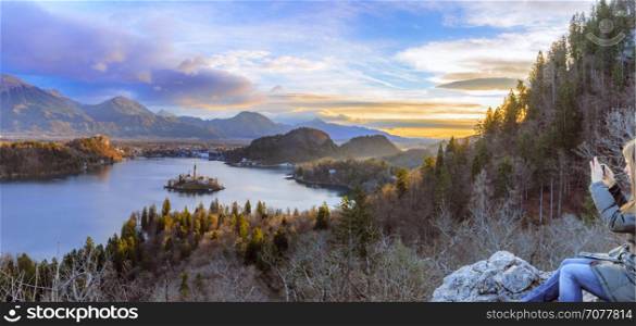 Panorama with the lake Bled under a colorful winter sky at sunrise and a young woman enjoys the view sitting on a rock