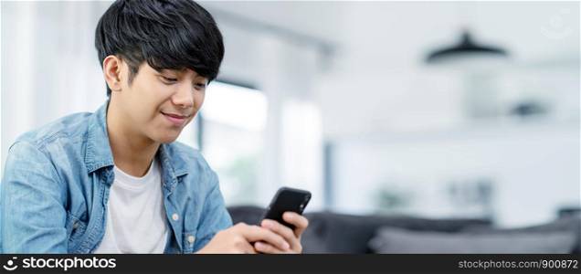 Panoramic or Banner of Happy asian teenager using smart phone and smiling on sofa living room at home. Asian man holding and using cellphone for searching data and social medie on internet.