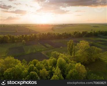 Panoramic view from the drone, aerial photography of landscape with greenery of forests, agricultural fields on the background of the cloudy sky at sunset in green and black colors.. Aerial view from the drone, a bird&rsquo;s eye view of abstract geometric forms of agricultural fields with a dirt road through them in the summer evening at sunset.