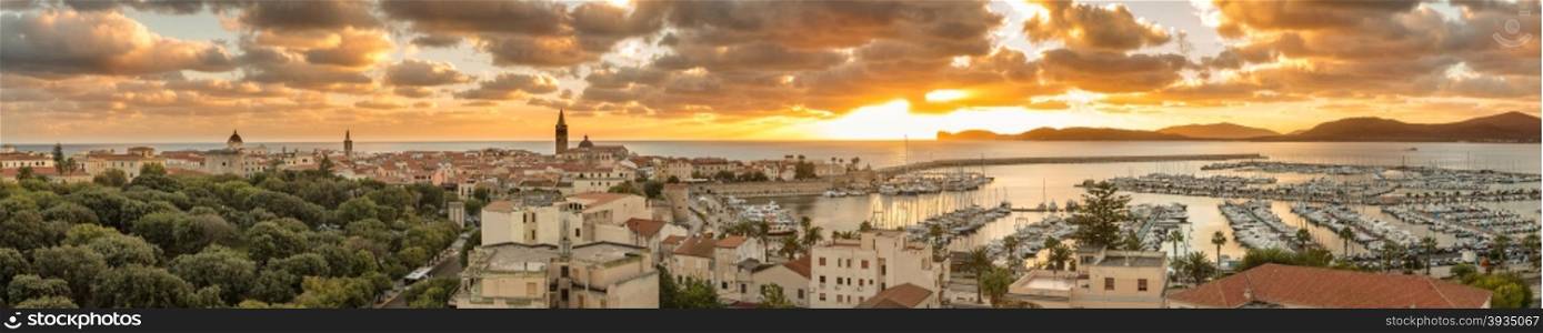 Panoramic view looking over the city and harbour of Alghero on the west coast of Sardinia with the sun setting behind the lighthouse at Capo Caccia