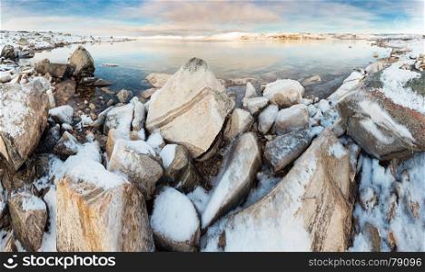 Panoramic view of norwegian lake at sunset, rocks covered with snow on foreground