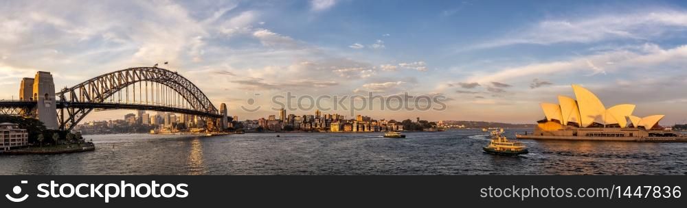 Panoramic view of Sydney harbor bridge and opera house with boats sailing in the bay and downtown in the background at sunset