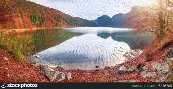 Panoramic view of the Alpsee lake, from Fussen, Germany, surrounded by mountains and forest in fall colors and reflected in its water.