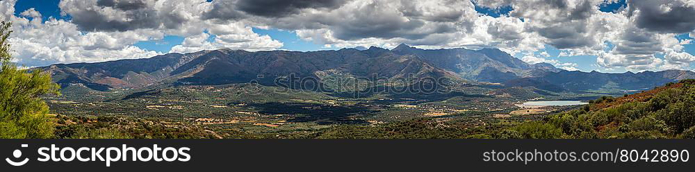 Panoramic view of the lush green Regino valley in the Balagne region of Corsica with mountains and mountain villages behind beneath a blue sky with fluffy white clouds and maquis in the foreground