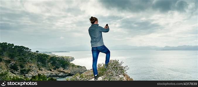 Panoramic view of young man standing on cliff photographing with smartphone, Alcudia, Majorca, Spain