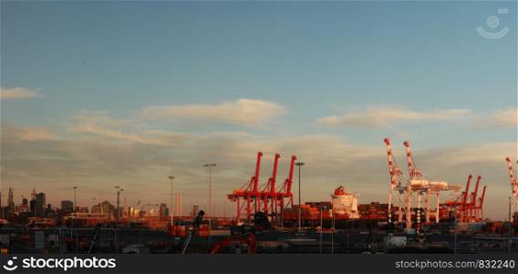 panoramic views of tall port shipping cranes standing tall loading a ship in port with shipping containers at Port Melbourne, with Melbourne city in the background, Victoria, Australia