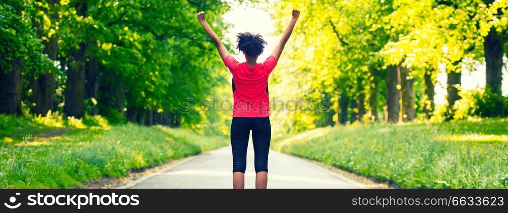 Panoramic web banner beautiful mixed race African American young woman girl teenager fitness running jogging celebrating success on road lined with spring or summer green trees