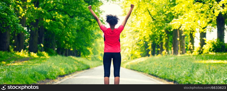 Panoramic web banner beautiful mixed race African American young woman girl teenager fitness running jogging celebrating success on road lined with spring or summer green trees