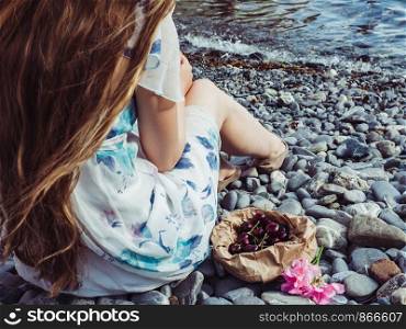 Paper bag with ripe cherries, a beautiful flower and a lonely girl with long hair against the background of a pebble beach and the sea. View from the back, close-up. Beautiful flower and a lonely girl with long hair