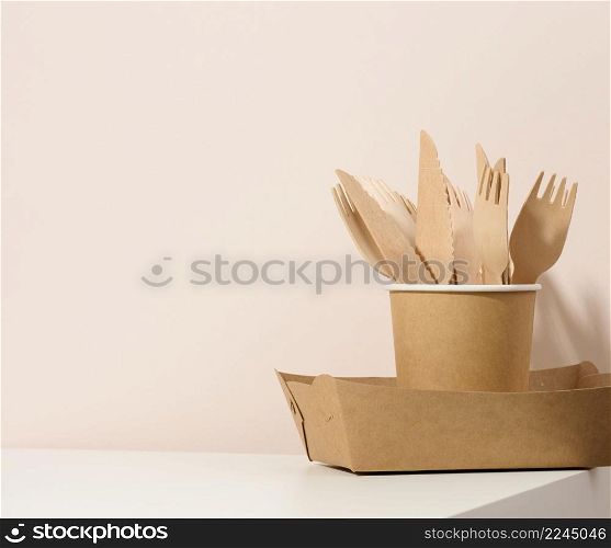 paper cardboard brown plates and cups, wooden forks and knives on a white table, beige background. Eco-friendly tableware, zero waste