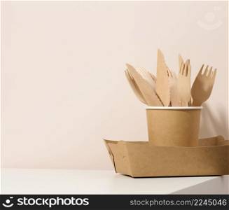 paper cardboard brown plates and cups, wooden forks and knives on a white table, beige background. Eco-friendly tableware, zero waste