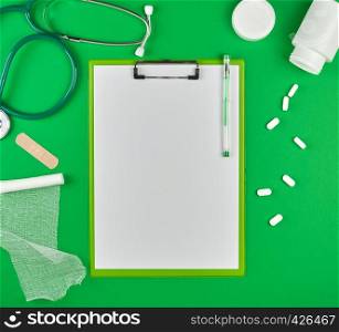 paper holder with empty white sheets, medical stethoscope, pills and textile bandage on a green background, top view, space for writing a prescription
