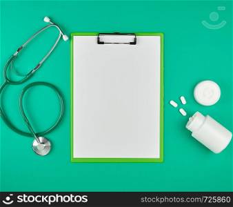 paper holder with empty white sheets, medical stethoscope, pills on a green background, top view, flat lay