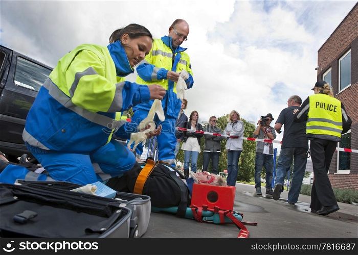 Paramedics tending to the first aid of an injured woman on a stretcher at the scene of a car crash, whilst a police woman is escorting a bystander towards the cordon tape, being filmed by a camera man