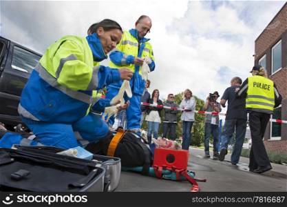 Paramedics tending to the first aid of an injured woman on a stretcher at the scene of a car crash, whilst a police woman is escorting a bystander towards the cordon tape, being filmed by a camera man