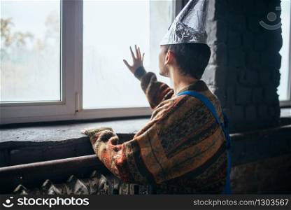 Paranoid man in tinfoil cap looking out the window, mind protection from telepathy, paranoia concept. UFO phobia, conspiracy theory