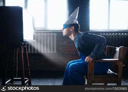 Paranoid man in tinfoil helmet and hypnotizing glasses watch TV, mind protection, paranoia concept. UFO, conspiracy theory, telepathy phobia