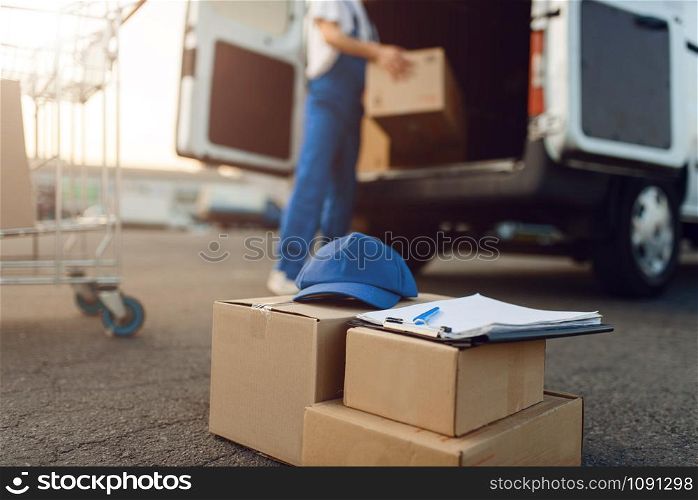 Parcel boxes and cap, deliveryman on background, delivery service. Man standing at cardboard packages in vehicle, male deliver, courier or shipping job