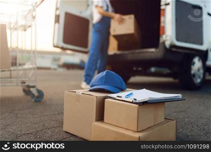 Parcel boxes and cap, deliveryman on background, delivery service. Man standing at cardboard packages in vehicle, male deliver, courier or shipping job