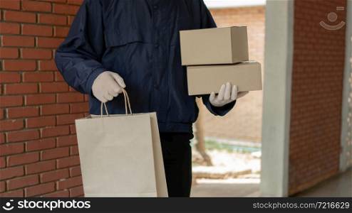 Parcel delivery concept the standing-tall post carrier who is wearing pale grey cap carrying one bag on left hand and two boxes on right hand and being ready to drop these stuffs to his customers.
